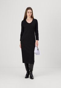 Vestido preto de manga longa com decote em V, feito de um tecido suave, que chega até a meia altura da panturrilha; combinado com botas pretas brilhantes até ao joelho e uma mala lilás clara.