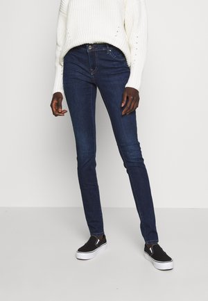 Personne portant un jean skinny bleu foncé, un pull en tricot blanc et des baskets slip-on noires avec des semelles blanches, debout devant un fond uni.