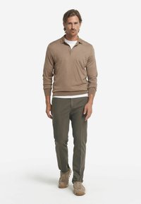 Brun stickad tröja med dragkedjekrage, bärs över en vit t-shirt. Olivgröna byxor och beige skor, står framför en enfärgad bakgrund.