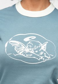 Simpel T-shirt i lyseblå med en hvid rund krave. Har et grafisk motiv af en sovende engel med teksten "WIP" på en fluffy skybaggrund.