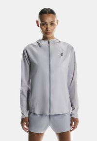 Svetlo siva jakna na zip s kapuco, izdelana iz gladkega, lahkega materiala. Ima logo na levem prsnem delu in prilagojen dizajn.