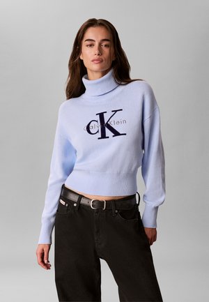 Femme portant un pull à col roulé bleu clair Calvin Klein et un pantalon noir, debout devant un fond gris uni.