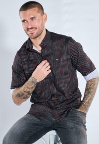 Schwarzes Hemd mit vertikalen roten und blauen Streifen, kurzen Ärmeln und einer geknöpften Vorderseite, kombiniert mit dunklen Jeans und einem hellen Unterhemd.