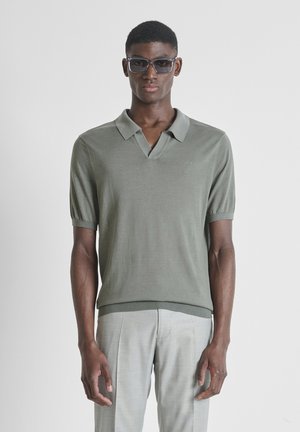 Modelo masculino con camisa gris de manga corta y cuello, pantalones gris claro y gafas de sol oscuras, de pie frente a un fondo gris liso.