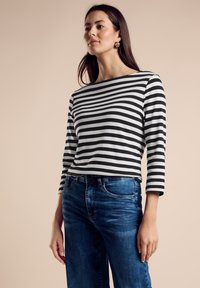 Czarna i biała, pasiasta koszula z długim rękawem; miękka tkanina; okrągły dekolt; noszona z niebieskimi dżinsami; model stojący na neutralnym tle.