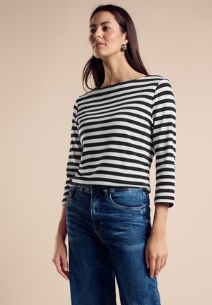 Gestreiftes Langarmhemd in Schwarz und Weiß; weicher Stoff; Rundhalsausschnitt; getragen mit blauen Denim-Jeans; Modell steht vor einem neutralen Hintergrund.