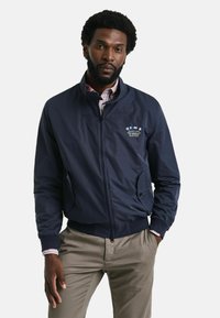 Marineblauwe bomberjack met een rits aan de voorkant, geribbelde manchetten en een logo op de borst. Ggedragen over een gestreefd shirt, met zijzakken.