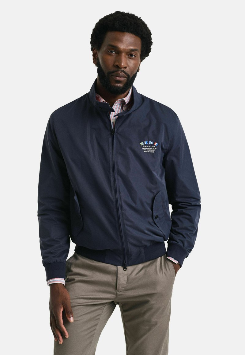 Marineblauwe bomberjack met een rits aan de voorkant, geribbelde manchetten en een logo op de borst. Ggedragen over een gestreefd shirt, met zijzakken.