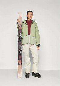 Ski ekipējums ietver gaiši zaļu jakku, sarkanu kreklu, bēšus bikses un melnus slēpošanas zābakus, ar slēpi modela rokā.