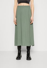 Longue jupe vert olive en tissu léger, dotée d'une taille élastique. Associée à des bottes noires montantes.