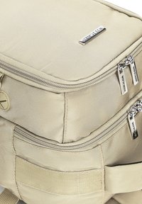 Beige textilní taška se dvěma stříbrnými zipy, značkovou visačkou, strukturovaným tvarem a texturovaným povrchem. Disponuje také dalšími kapsami a popruhy.