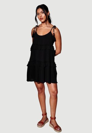 Frau trägt ein ärmelloses, mehrlagiges schwarzes Kleid mit Bindeträgern und braune Keilsandalen, steht vor einem schlichten hellgrauen Hintergrund.