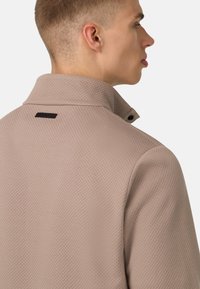 Giacca beige con colletto in tessuto testurizzato, dotata di un alto colletto e un'etichetta con logo nero sul retro. Dettaglio semplice con bottone al colletto.