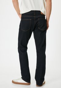Jeans de color azul oscuro con corte recto, que presentan costuras naranjas y bolsillos traseros, combinados con zapatillas blancas sobre un fondo liso.
