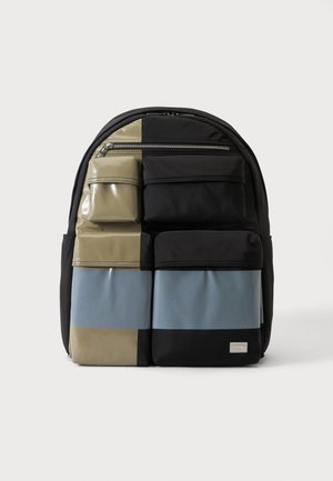 Sac à dos avec un design à blocs de couleur noir, beige et bleu, comportant plusieurs poches avant zippées et compartiments.