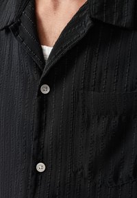 Sort button-up skjorte med et tekstureret vertikalt stribemønster, en lomme og lyse knapper. Stoffet ser blødt og glat ud.