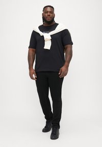 Μαύρη μπλούζα T-shirt με λογότυπο, μαύρο στενό παντελόνι και μαύρα αθλητικά παπούτσια. Ένα κρεμ πουλόβερ είναι ριγμένο χαλαρά στους ώμους.