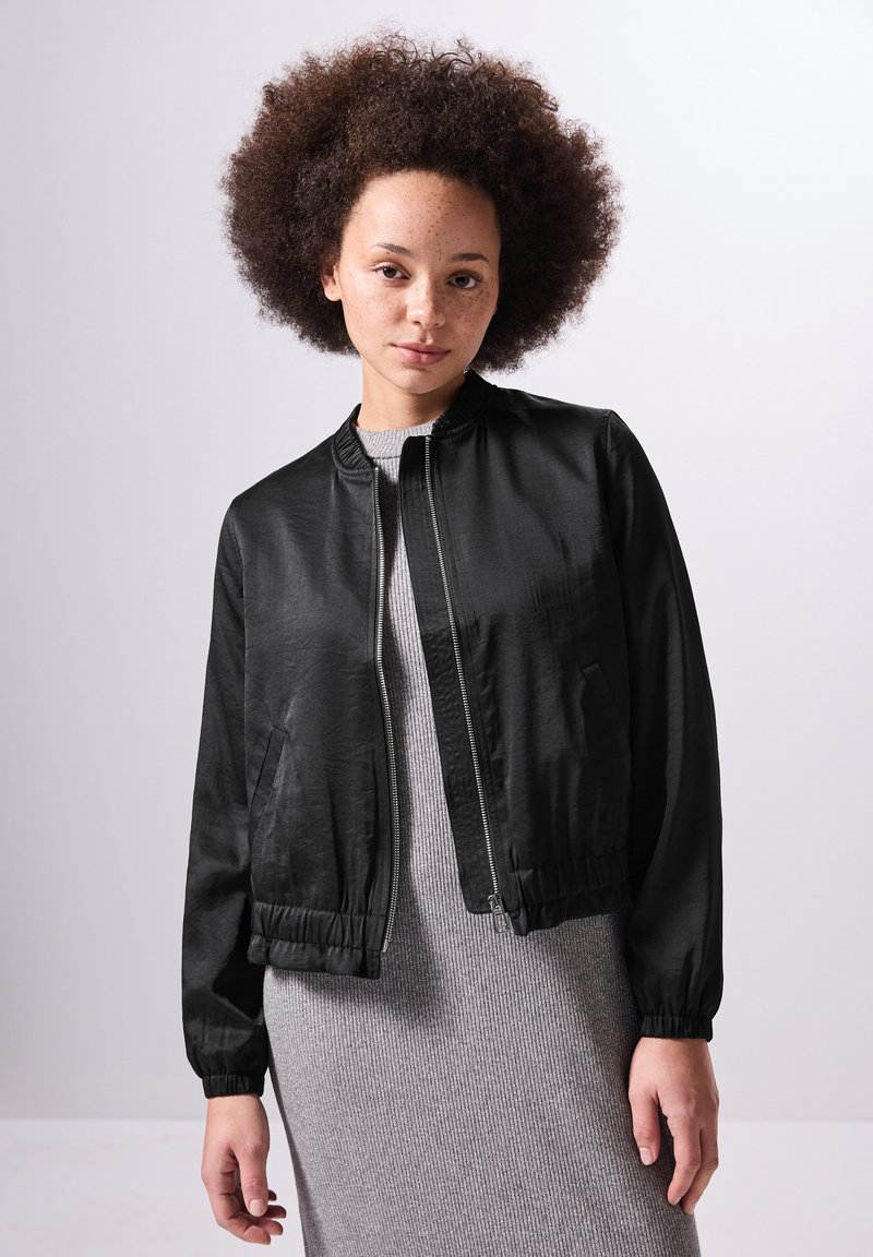 Veste bomber noire avec fermeture éclair, poignets et ourlet élastiqués. Portée sur une robe grise à côtes, mise en valeur par une texture lisse et brillante.