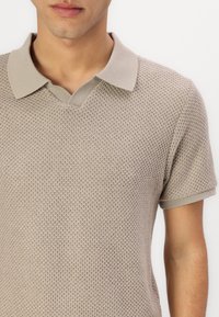 Beige kötött poloing, texturált mintával, V-nyakkal, és kontrasztos világos bézs gallérral és ujjaikkal. Az anyag légáteresztőnek tűnik.