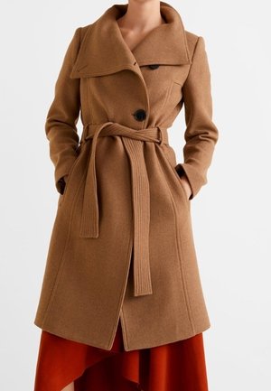 Femme portant un manteau en laine marron longueur genou avec un large col, des boutons noirs et une ceinture nouée, sur une jupe ou une robe asymétrique rouge.