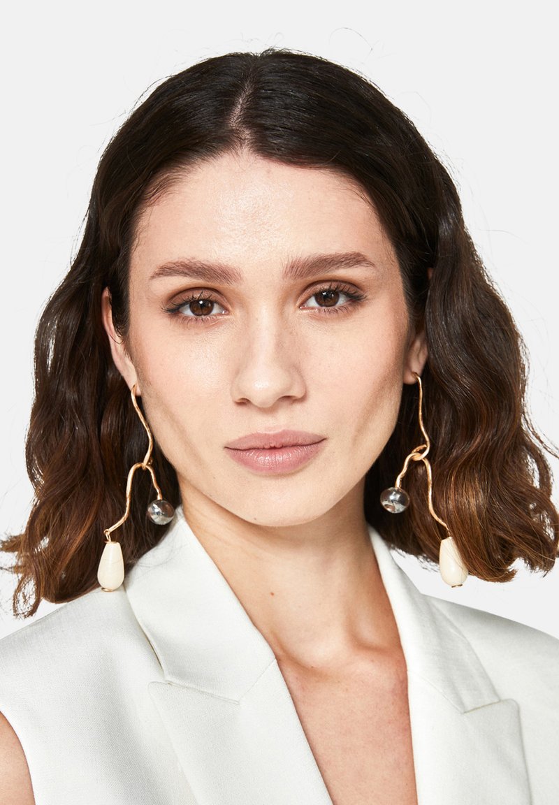 Guldtonade örhängen med en vågig design, iögonfallande klara pärlor och en elfenbensfärgad droppformad detalj. Slät yta med en polerad finish.