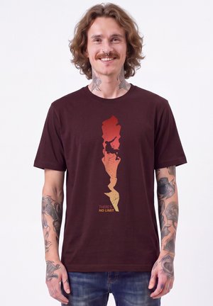 Homme souriant aux cheveux mi-longs et aux tatouages porte une chemise bordeaux montrant la silhouette d’un grimpeur sur un rocher avec le texte "There's no limit."