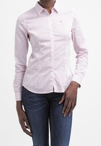 Rood en wit gestreepte lange mouwen button-up shirt, met een kraag, knopen aan de voorkant en een klein logo op de borst. Gecombineerd met donkere jeans.