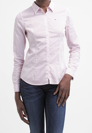 Red and white striped long-sleeve button-up shirt, featuring a collar, front buttons, and a small logo on the chest. Paired with dark jeans.