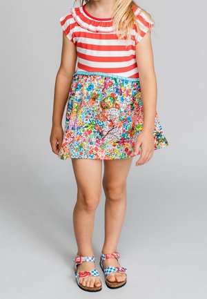 Czerwono-biała bluzka w paski z falbaniastym dekoltem, zestawiona z kolorową spódnicą w kwiatowy wzór. Sandały z niebieskimi akcentami z czerwonymi kwiatami.