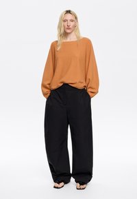Svetlo rjav oversized pulover z dolgimi rokavi, v kombinaciji z ohlapnimi črnimi hlačami z bočnimi žepi in gladko teksturo.