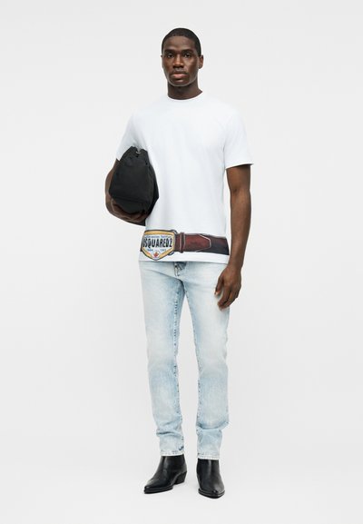 Άνδρας στέκεται κρατώντας μαύρη τσάντα, φοράει λευκό t-shirt με τύπωμα ζώνης, ανοιχτόχρωμα μπλε τζιν και μαύρα μποτάκια σε απλό ανοιχτό φόντο.