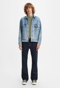 Light blue denim jacket over a green t-shirt, paired with dark jeans and black sneakers. Eleven visible buttons on the jacket, relaxed fit.