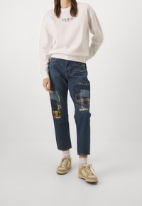 Vit sweatshirt med "POLO" logotyp, blå denimjeans med lapptäckesdetaljer i rutigt och olika nyanser. Beiga sneakers och vita strumpor.