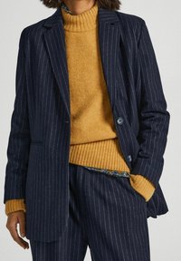Woman wearing navy blue pinstripe blazer and pants with mustard yellow ribbed turtleneck sweater, one hand in pocket, against plain background.