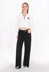 Hoodie bianco zipato tagliato con grafica di labbra rosse, abbinato a pantaloni neri a gamba larga a vita alta e sneakers nere con suola bianca.