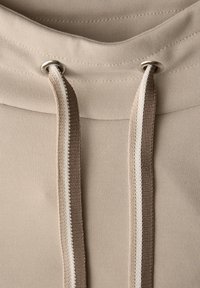 Detalle de sudadera con capucha beige con una textura suave, que presenta dos cordones — uno beige y otro marrón — fijados con ojales plateados.