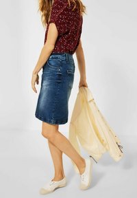 Jeanskjol i mörkblått med en figursydd silhuett och lätt slitning, matchad med en mönstrad top i vinröd och vita sneakers. En ljusgul jacka hänger.