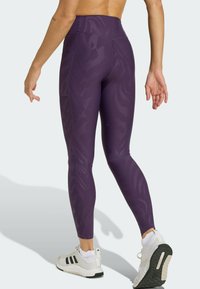 Lila Leggings mit hoher Taille und subtiler Ton-in-Ton-Struktur, aus elastischem Stoff, kombiniert mit weißen Sportschuhen mit schwarzen Akzenten.