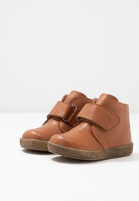 Bottines en cuir marron avec une sangle Velcro et une semelle en caoutchouc texturée, présentant des coutures simples et un design à bout rond.