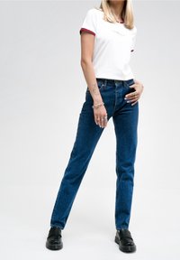 Camiseta blanca con ribete rojo y texto, combinada con jeans de un azul oscuro. El modelo lleva zapatos negros sin cordones, mostrando un atuendo casual.
