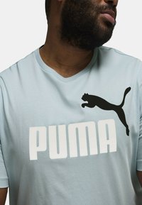 Camisa de manga corta azul claro hecha de tela suave, con un gran logo blanco de "PUMA" y un gráfico de un puma saltando en negro en el pecho.