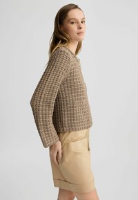 Beige strukturierte Strickjacke mit langen Ärmeln, rundem Ausschnitt, Knopfverschluss und zwei frontalen Taschen, kombiniert mit beigen Shorts.
