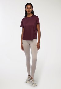 Bordowy, krótki top sportowy z krótkim rękawem w połączeniu z beżowymi legginsami i szarymi sneakersami. Gładki materiał o luźnym kroju z detalem logo na dolnym brzegu.