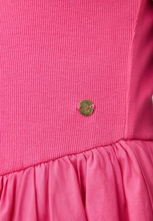 Ružový top z prúžkovaného materiálu so zhrnutým lemom a malou zlatou kovovou značkou v tvare kruhu s vyrytým písmenom "B" pripevnenou v páse.