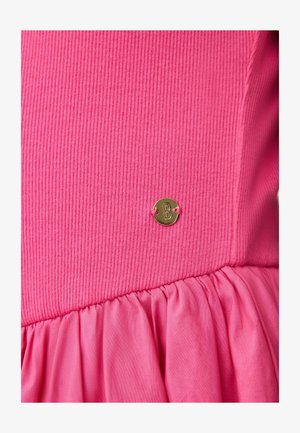 Rožinis ribuotas audinio viršus su surinktą apačia ir mažu apvaliu auksiniu metalo ženkliuku su įrašu „B“, prisegtu prie liemens.