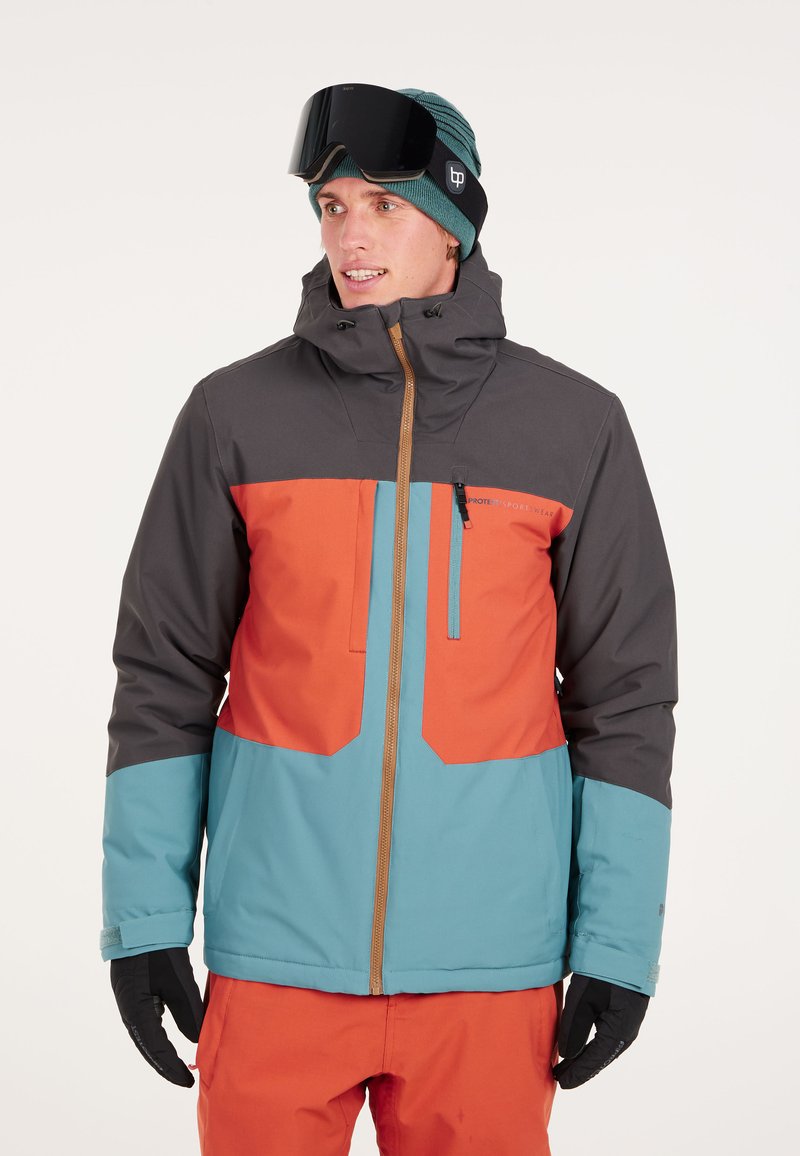 Hombre con chaqueta de invierno en color verde azulado, naranja y gris, pantalones naranjas, gorro verde azulado, gafas de esquí negras y guantes negros, mirando hacia la derecha.