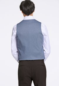 Blå texturerad väst med en skräddarsydd passform, bärs över en vit skjorta, i kombination med mörka byxor. Har en låg rygg och ren fåll.