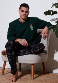 Sötétzöld, hosszú ujjú póló, ami egy rajzfilmmedvét ábrázol, fekete kockás pizsamanadrággal párosítva. Egy világos színű széken ül.