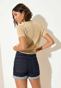 Beiges, kurzärmliges, gestricktes Oberteil mit geripptem Saum, kombiniert mit dunkelblauen Jeans-Shorts, die am Saum aufgerollt sind. Minimalistisches Design, keine sichtbaren Muster oder Akzente.