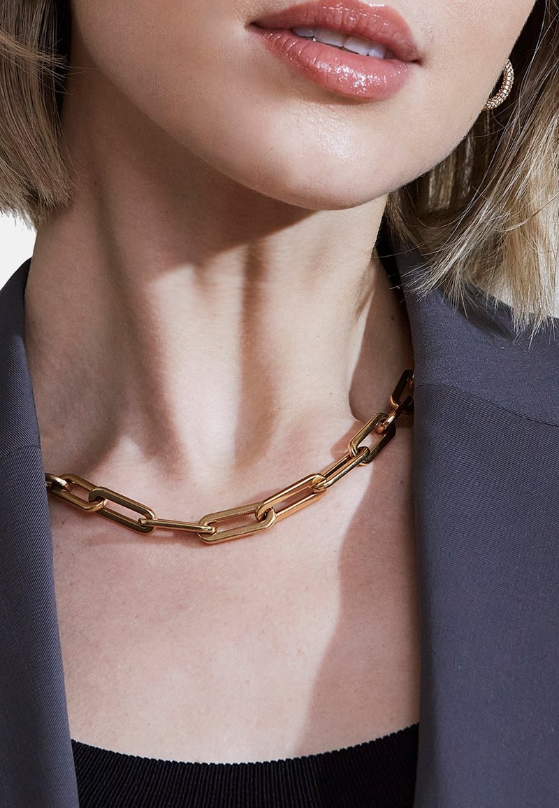 Collana in metallo dorato con catena caratterizzata da anelli allungati, superficie liscia e design semplice, indossata con un blazer grigio.